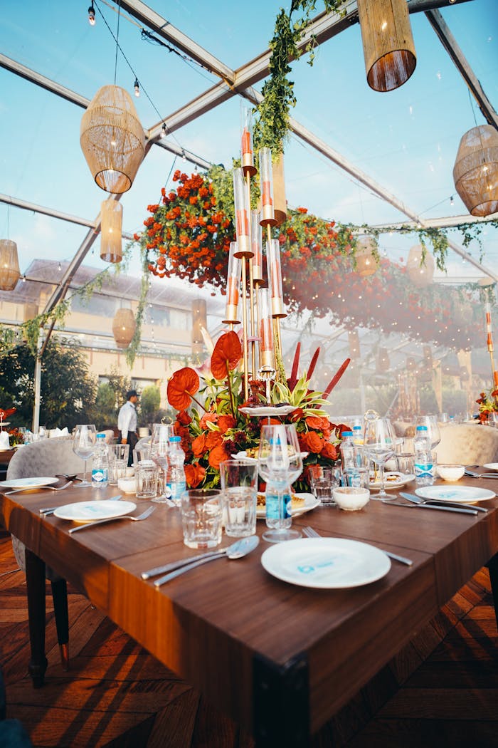 Luxurious table setting for a wedding under a transparent tent with floral arrangements and hanging lights.